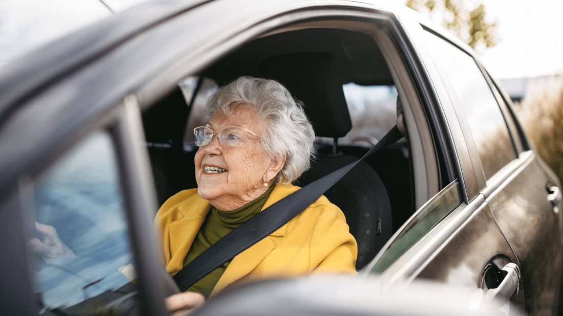 An elderly woman in a car during her driving test, smiling and relaxed.